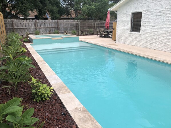 Piscina retangular com borda azulejada, rodeada por um jardim com diversas plantas e um cercado de madeira ao fundo.