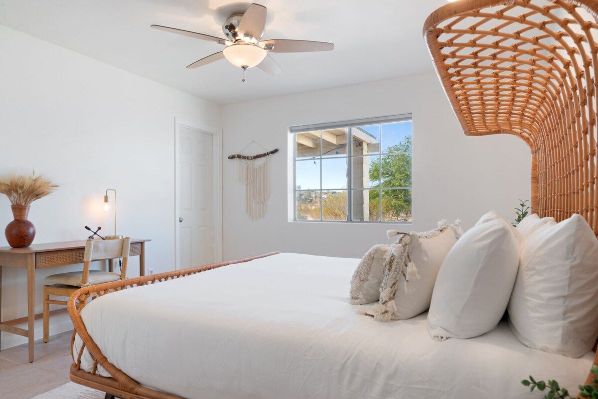 A bedroom with a large bed, a desk, a chair, a ceiling fan, and a window with a view.