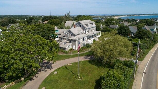 A large, two-story house with a prominent American flag, surrounded by lush greenery and a clear view of the ocean.