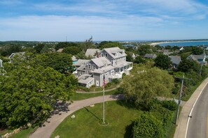 A large, two-story house with a prominent American flag, surrounded by lush greenery and a clear view of the ocean.