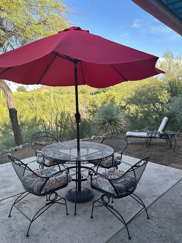 An outdoor patio with a round metal table and chairs, a red umbrella, and a view of a landscaped garden.