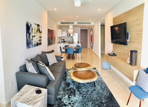 Un salon moderne avec un canapé gris, une table basse et un fauteuil bleu. On aperçoit une cuisine à l’arrière-plan, avec un coin repas.