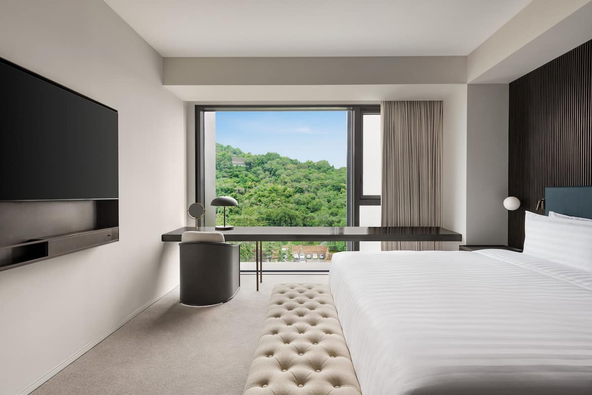 Een moderne hotelkamer met een groot bed, een flatscreen-tv aan de muur, een klein bureau met een lamp en uitzicht op een groen landschap door een groot raam.