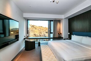 Una habitación de hotel moderna con una cama grande, televisión, balcón con vista a la ciudad y un amplio ventanal.