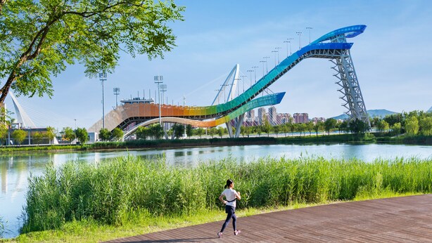Un corredor en una passera de fusta al costat d'un riu, amb un pont modern de fons.