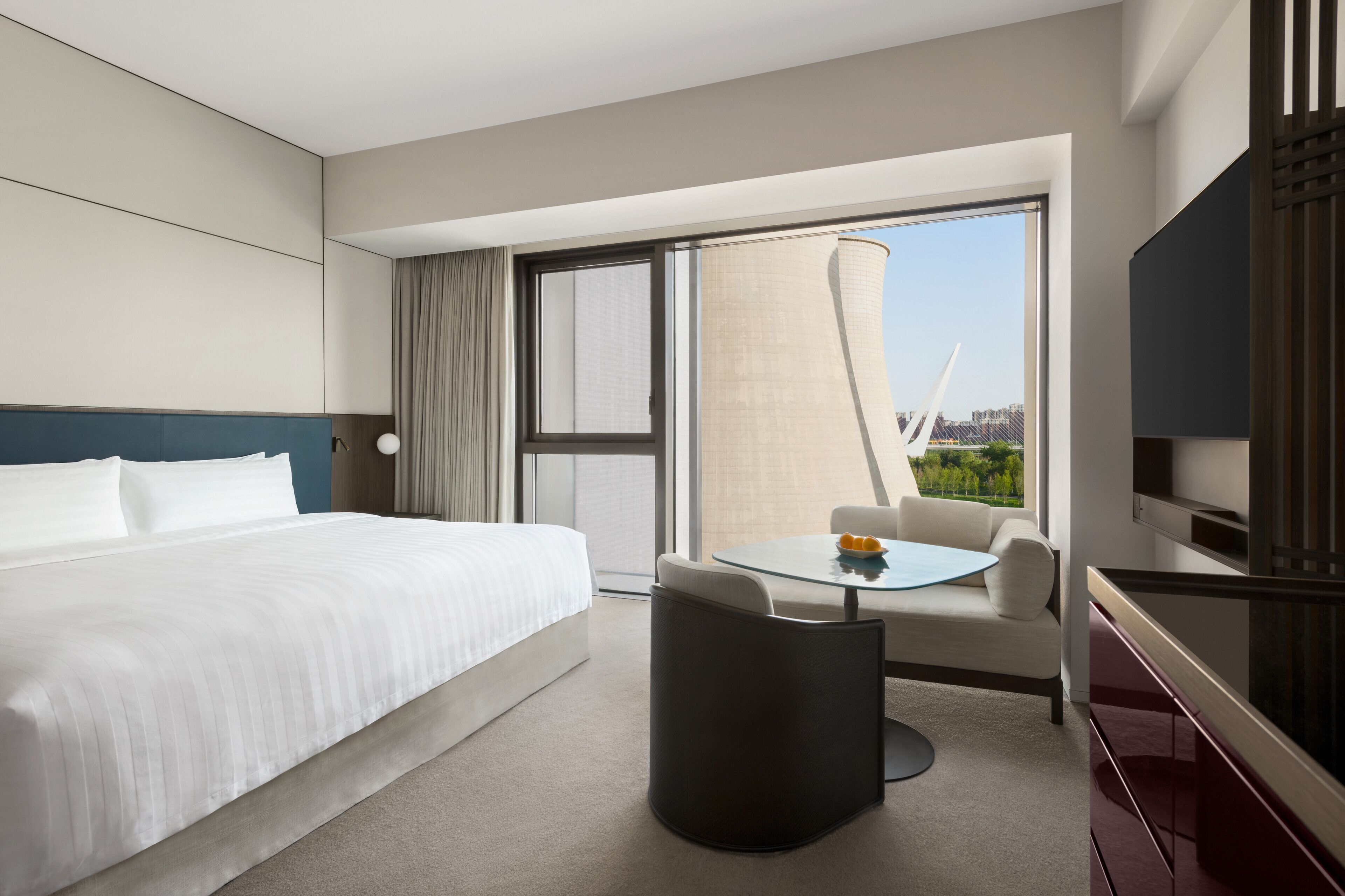 Een hotelkamer met een bed, een klein tafeltje met een stoel, een televisie en een groot raam met uitzicht op gebouwen.