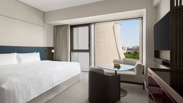 Een hotelkamer met een bed, een klein tafeltje met een stoel, een televisie en een groot raam met uitzicht op gebouwen.