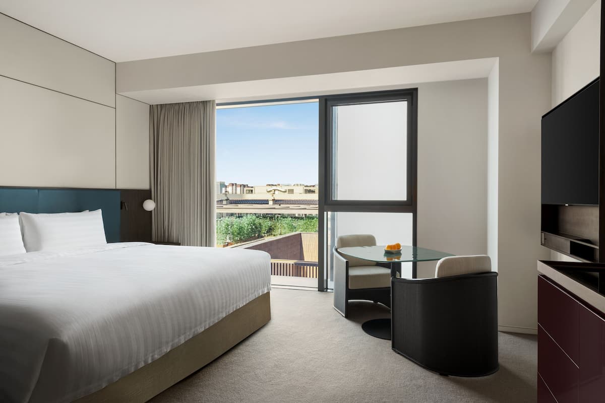 Hotelkamer met een groot bed, een klein tafeltje met een stoel, een televisie en een balkon met uitzicht op gebouwen en groen.
