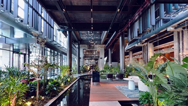 Un espacio interior moderno con una fuente central, zonas de estar y abundante vegetación de interior.