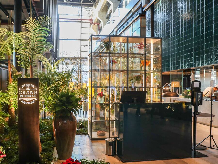 A modern indoor space with a glass enclosure displaying plants and flowers, a wooden sign, and a kitchen area with a chef.