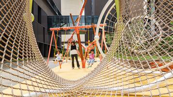 Un parque infantil con estructuras de escalada cubiertas por redes y columpios, frente a la fachada de un edificio moderno.
