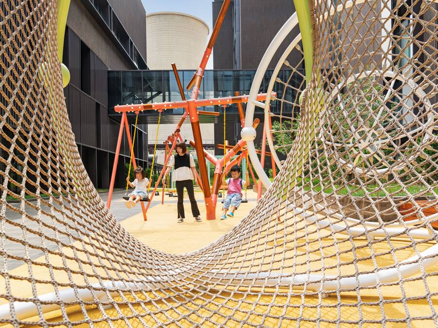 Un parque infantil con estructuras de escalada cubiertas por redes y columpios, frente a la fachada de un edificio moderno.