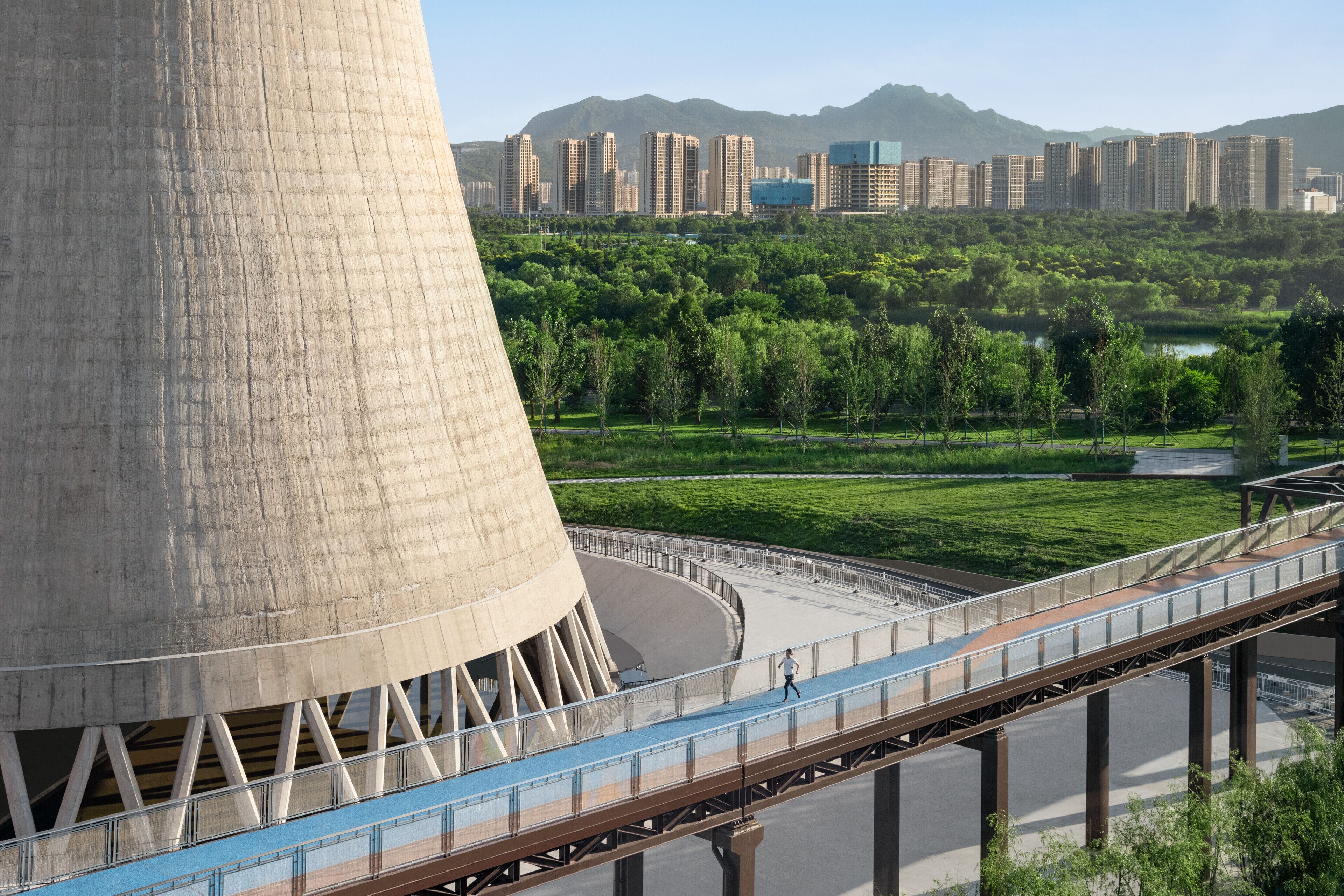 A large industrial structure with a curved base, a pedestrian bridge, and a cityscape with high-rise buildings in the background.