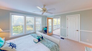 A bedroom with a bed, a ceiling fan, large windows, a door, and a view of the outdoors.