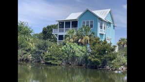 A two-story house with a balcony, surrounded by dense vegetation and a body of water.
