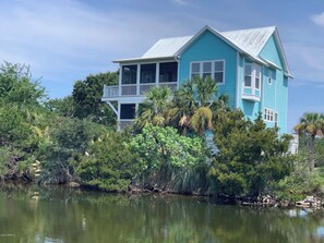 A two-story house with a balcony, surrounded by dense vegetation and a body of water. - Extended Golf cart! Elevator! Amazing Views! Pets! Accessible! 4 decks! (Saint Helena Island)