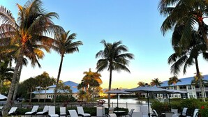 Eine Resort-Terrasse mit Liegestühlen und Sonnenschirmen, Palmen und Blick auf ein Gebäude mit blauem Dach.
