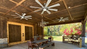 A covered outdoor deck with wooden flooring, a central table with chairs, and two ceiling fans.