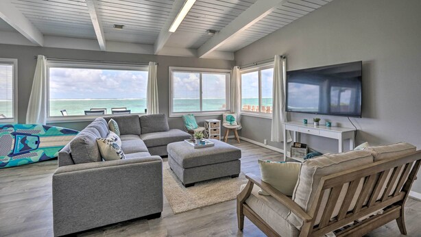 A spacious living room with a large TV, grey sofas, a wooden chair, and a view of the ocean through large windows. - WFH in Paradise: Beachfront Hauula Getaway! (Hauula)