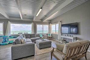 Un salón amplio con un televisor grande, sofás grises, una silla de madera y vistas al océano a través de ventanas grandes.