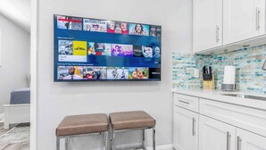 A modern kitchen with a large TV displaying various movie and TV shows, two bar stools, and a tiled backsplash. - Kick back and relax in this calm, stylish space (Fort Lauderdale)
