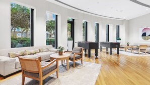 Un salon spacieux comprenant un canapé, des fauteuils, une table basse et deux tables de baby-foot. De grandes fenêtres laissent entrer la lumière naturelle et offrent une vue sur la verdure environnante.