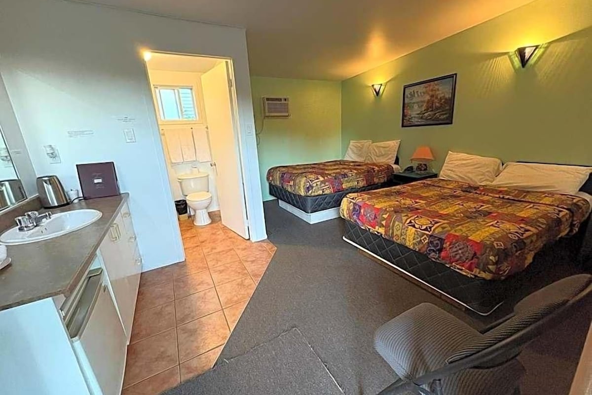 A room with two beds, a sink, and a chair. There is a door leading to a bathroom.
