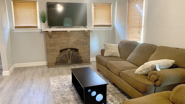 Una sala de estar con una televisión de pantalla plana sobre una chimenea de ladrillo, un sofá marrón con cojines, una mesa de centro negra y persianas de madera en las ventanas.