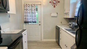 Una cocina compacta con armarios blancos, un refrigerador negro, un microondas y un lavavajillas. Una cortina estampada cubre una puerta, y se observa un reloj de pared decorativo.