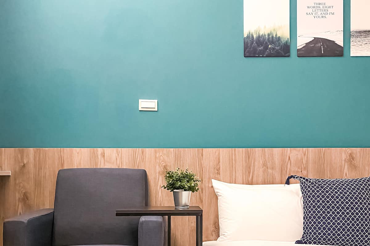 A room with teal walls, a wooden headboard, a grey armchair, a small table with a plant, and a bed with white and patterned pillows.