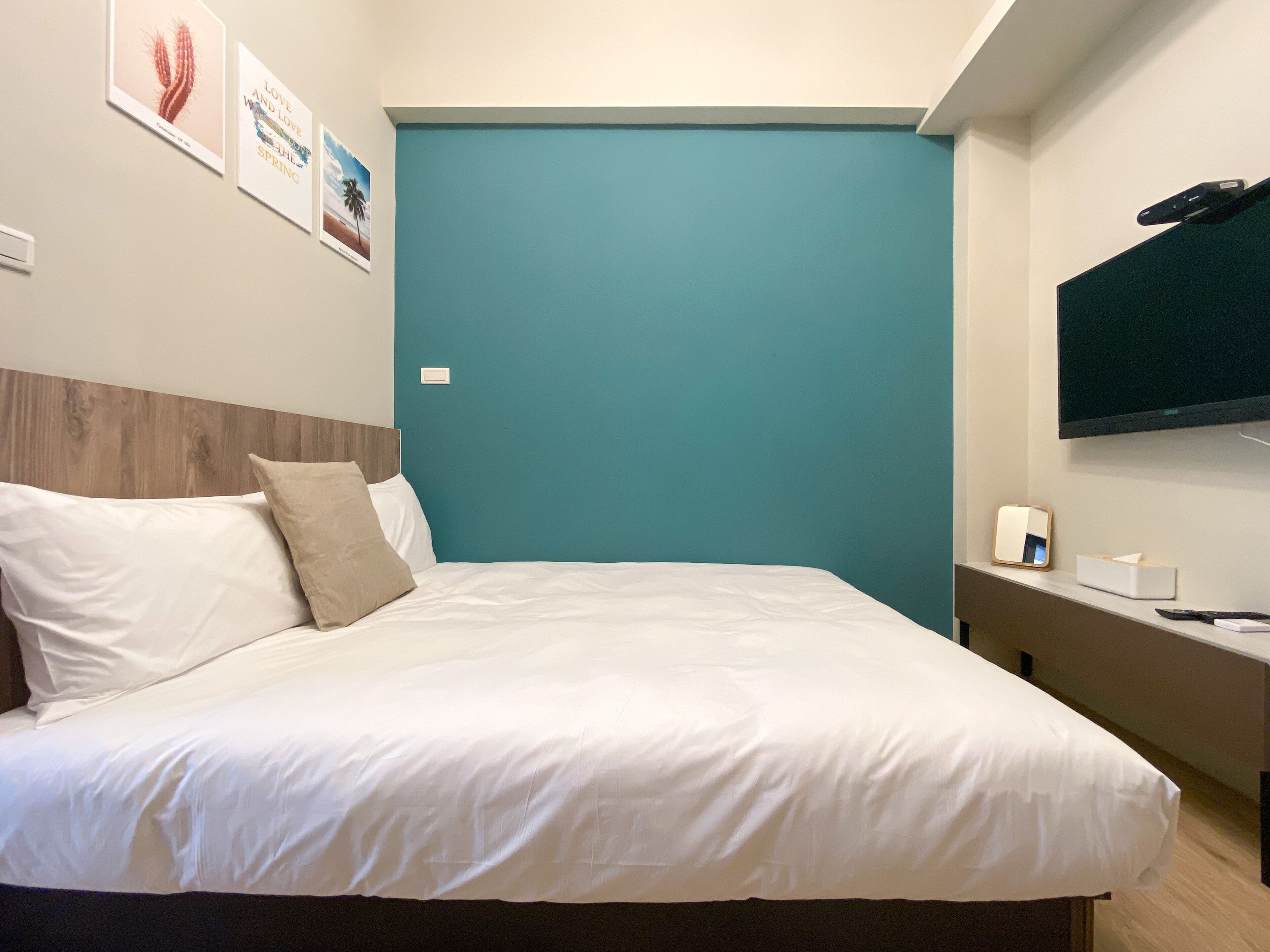 A neatly made bed with white linens and beige pillows, a teal accent wall, a mounted flat-screen TV, and a bedside table with a remote control and a tissue box.