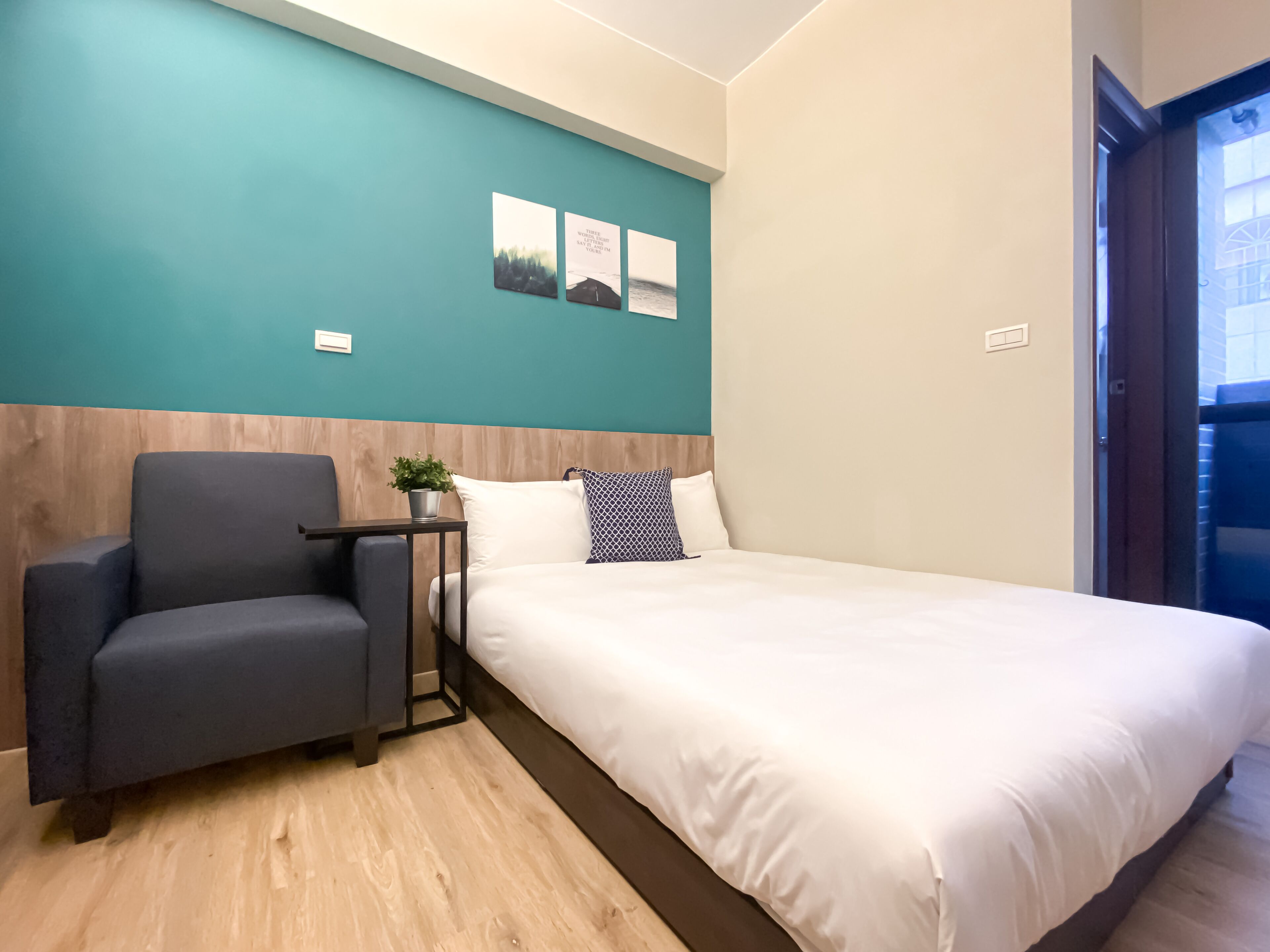Habitación de hotel con cama, una silla, una mesita con una planta y tres cuadros en la pared azul turquesa.