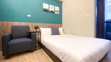 Habitación de hotel con cama, una silla, una mesita con una planta y tres cuadros en la pared azul turquesa.