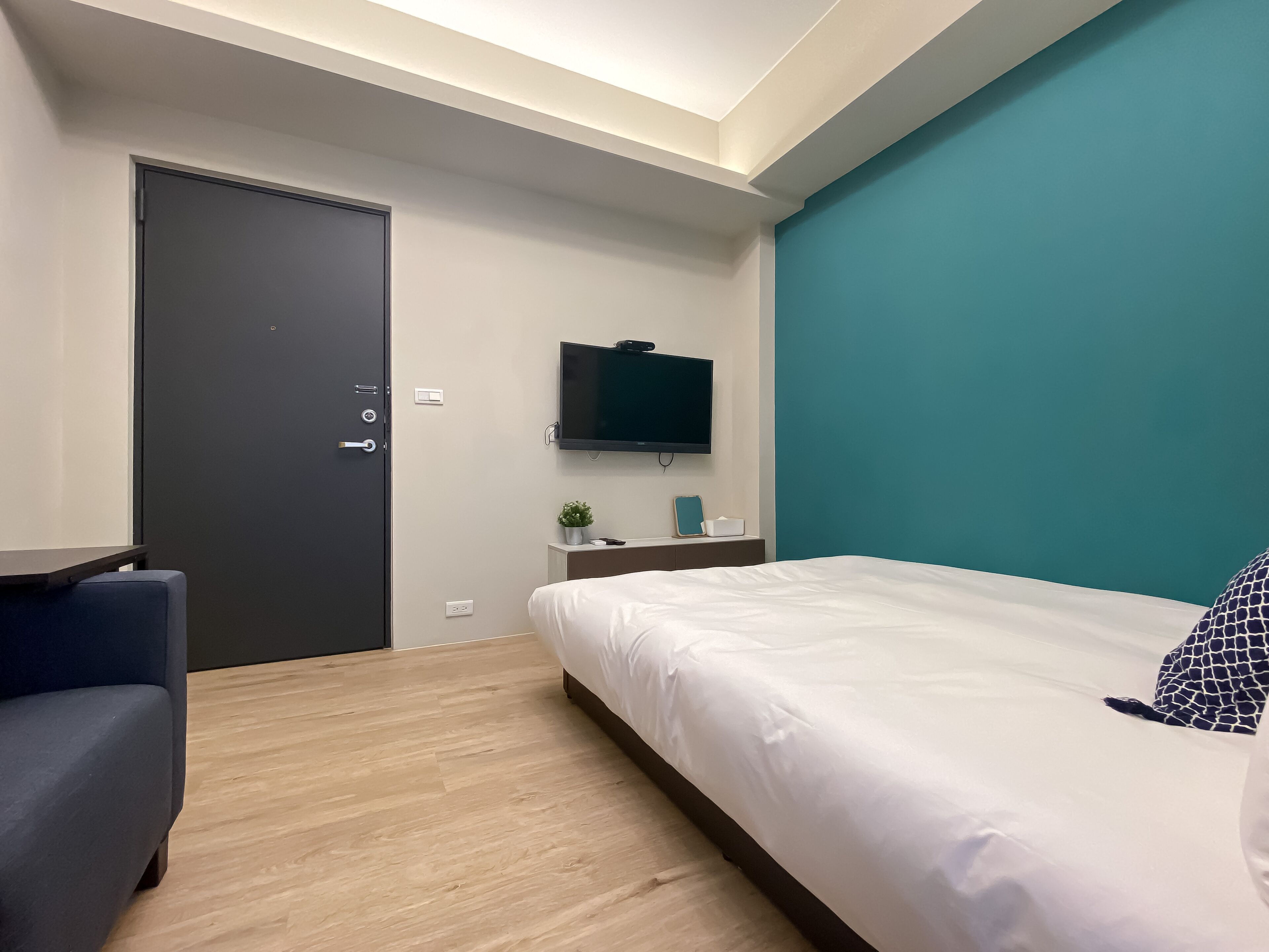 A modern hotel room with a bed, a desk, a chair, a TV, and a door.
