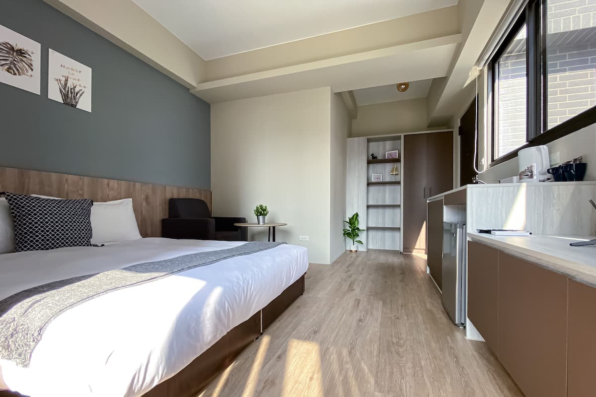 A modern hotel room with a large bed, a desk, a chair, and a view of the kitchen area.