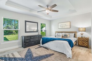 A bedroom with a bed, dresser, nightstand, ceiling fan, and large windows.