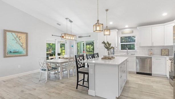 Una cocina moderna con armarios blancos, una isla central, iluminación colgante y un comedor con sillas y una mesa blancos.