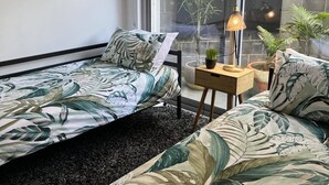 Quarto com duas camas, uma mesa pequena com abajur e vista para um jardim através de janelas amplas.