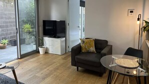 Uma sala de estar moderna com uma mesa de vidro, sofá, televisão e uma porta de correr de vidro que dá para uma varanda com plantas.