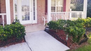 A brick house with a white porch, a white door, and a small garden with bushes and a metal fence. - Welcome to Eve's Place. Come enjoy the historic township of Conyers. (Conyers)