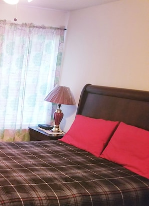 Una cama tamaño queen con cubrecama de cuadros y almohadas rojas, una lámpara de noche y una ventana con cortinas translúcidas.
