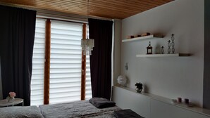A bedroom with a wooden ceiling, a bed with patterned bedding, a chandelier, and a window with blinds.