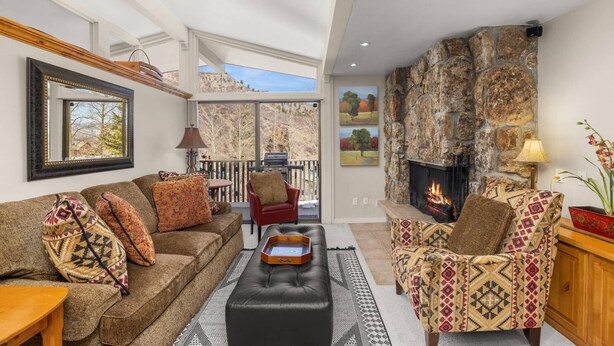 A cozy living room with a fireplace, a large window, and a stone accent wall. - Popular Snowmass Condo with HT/Wood FP/Balcony W/Grill/Parking. Great Rates! Plan Your Summer Now! (Snowmass Village)