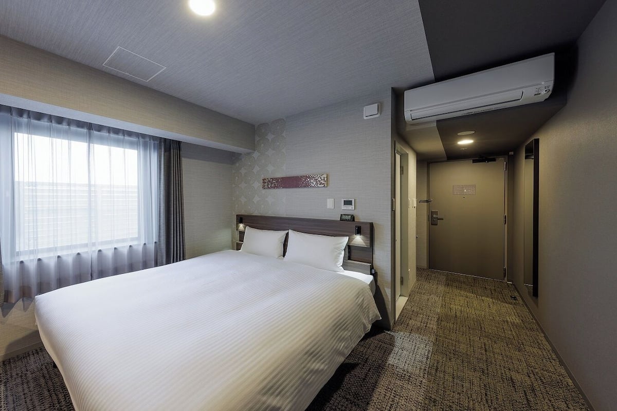 A hotel room with a large bed, a window with curtains, an air conditioning unit, a wall clock, and a carpeted floor.