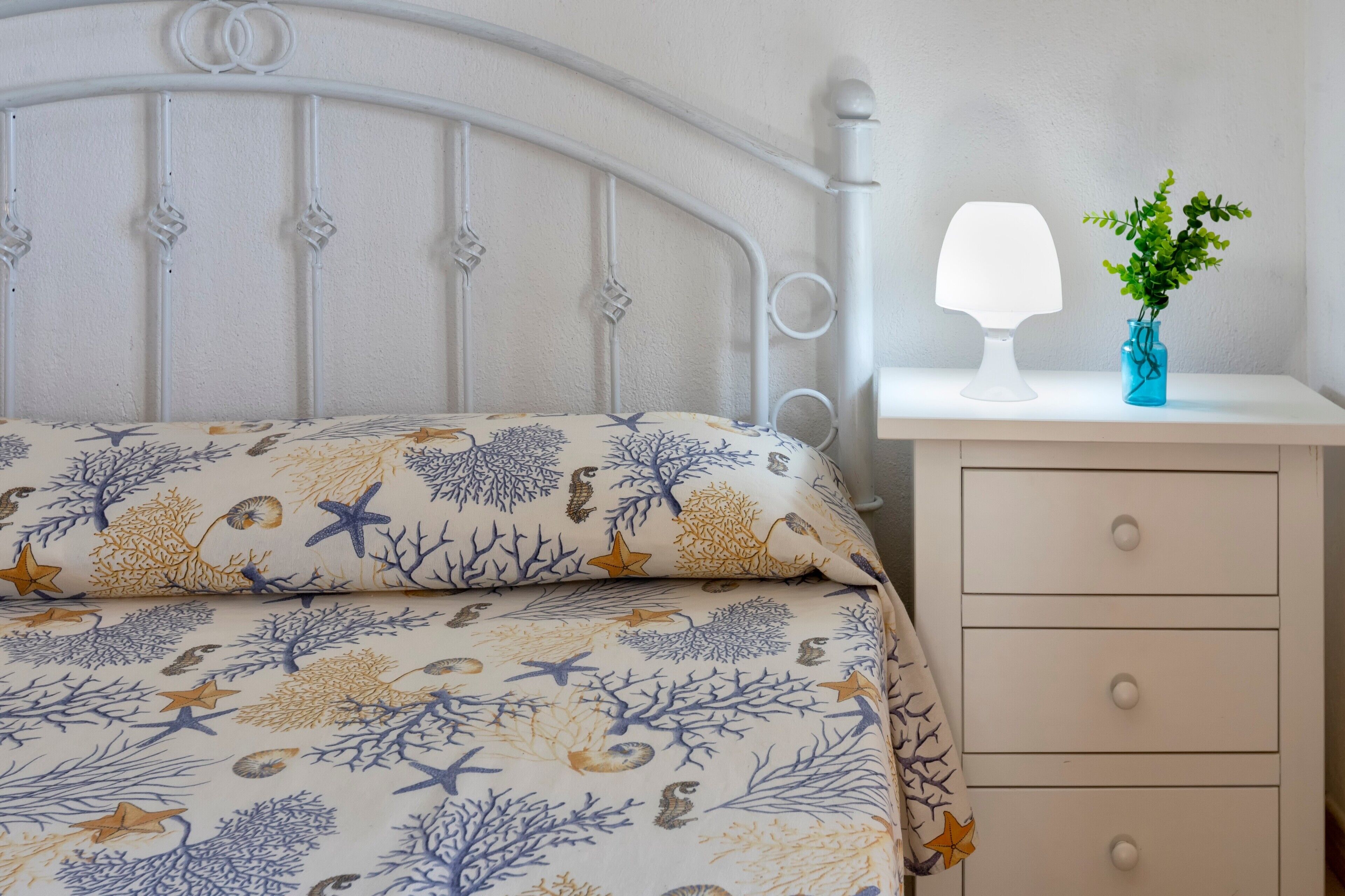 Un lit avec une couvre-lit à motifs, une table de chevet avec une lampe et un vase, et une commode.