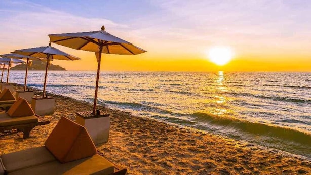 Een strand met ligbedden en parasols, een heldere lucht, en de zon die in de oceaan ondergaat.