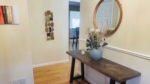 A hallway with a wooden floor, a dark wooden console table with a vase of flowers, a wall-mounted mirror, and a geometric wall art piece.