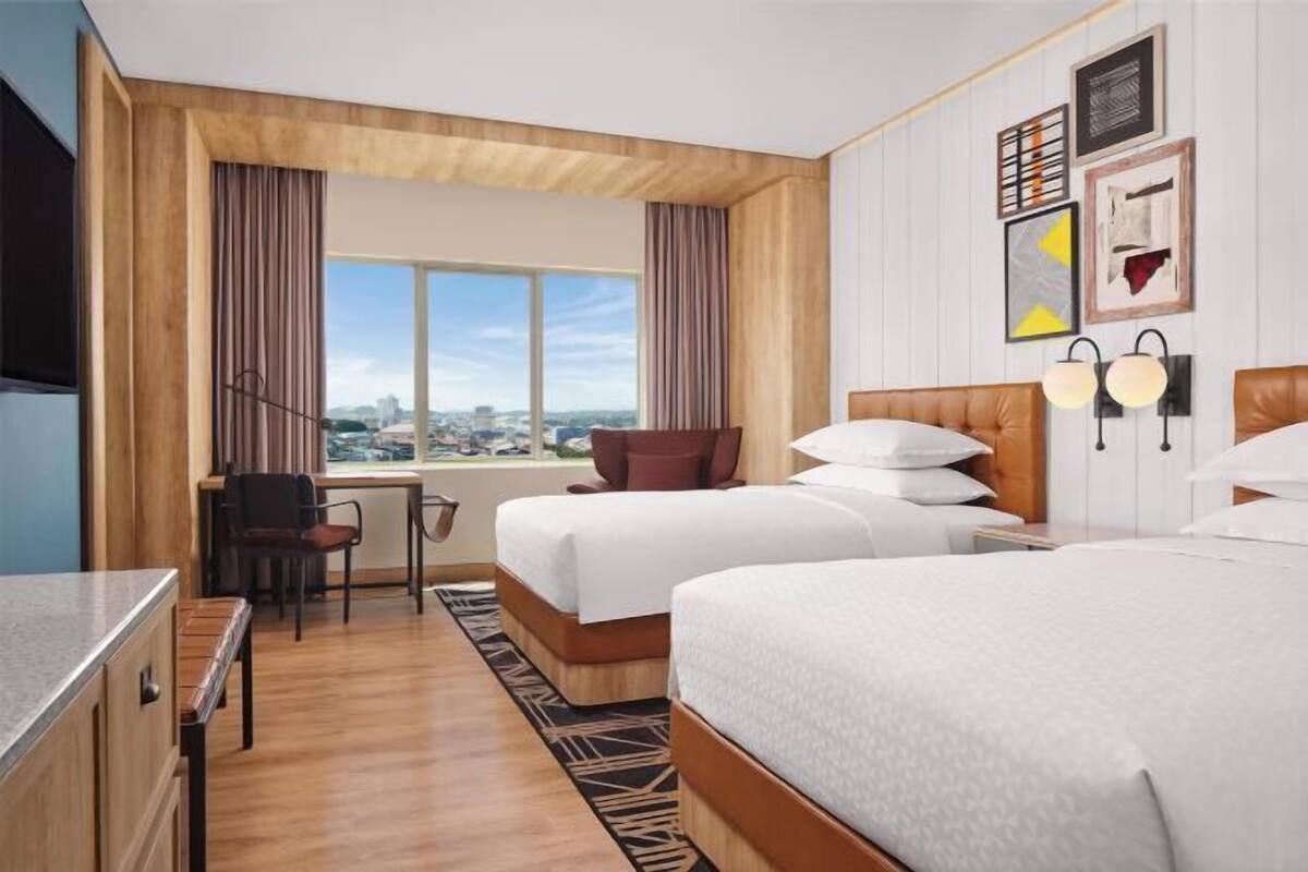 A hotel room with two beds, a desk, a chair, a window with a city view, and wall-mounted lights.