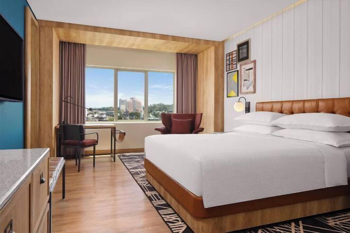 A hotel room with a bed, a desk, a chair, a window with a city view, and a wall with framed pictures.
