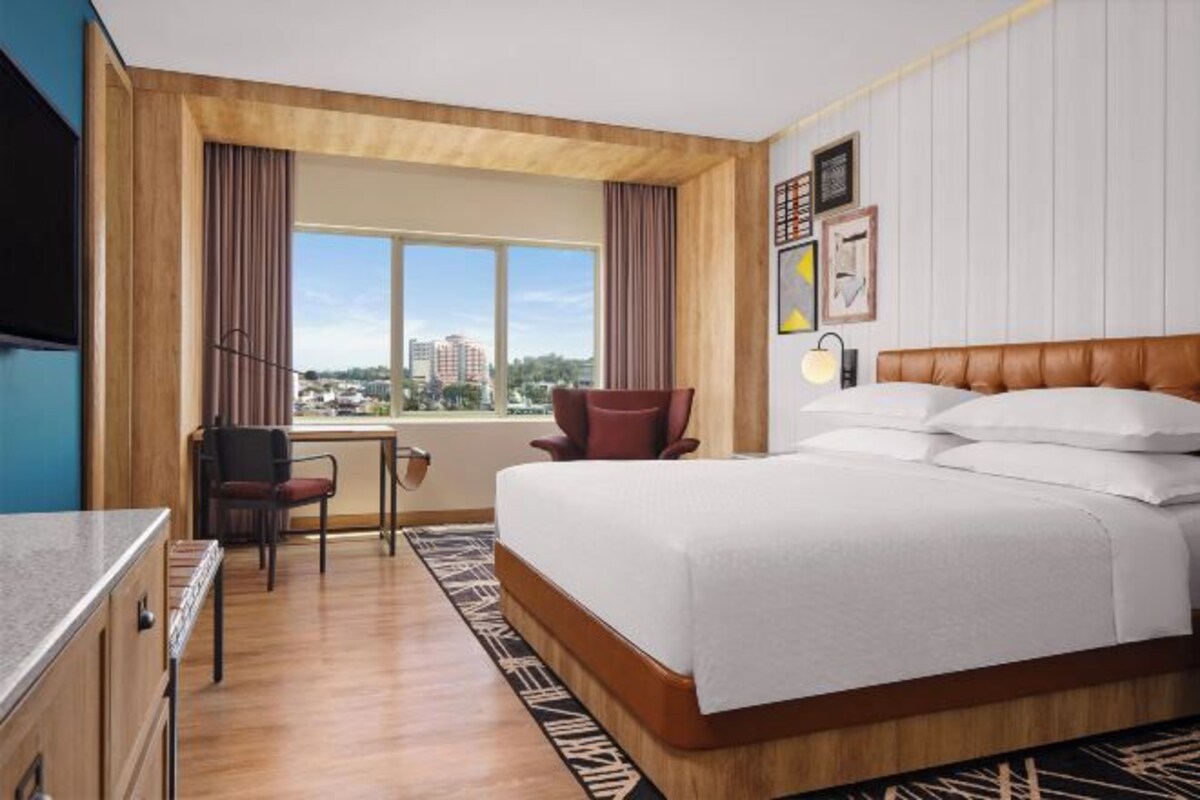 Ein Hotelzimmer mit einem Bett, einem Schreibtisch, einem Stuhl, einem Fenster mit Stadtblick und einer Wand mit gerahmten Bildern.